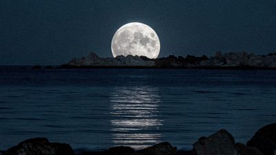 Полная Луна в ясном ночном небе над спокойным морем у скалистого берега