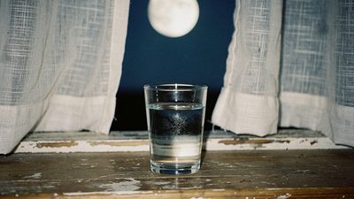 Тёмная комната с одной свечой и стаканом воды у окна, в окне видна луна над городскими крышами
