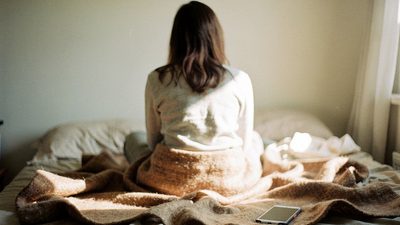 Женщина со спины на краю кровати в светлой комнате утром, телефон рядом на пледе