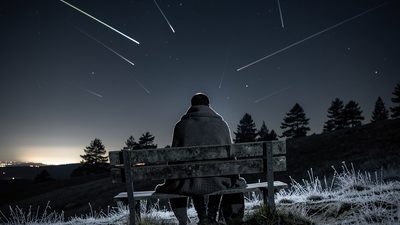 Силуэт человека в пледе на скамейке под тёмным небом со следами метеоров над соснами