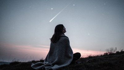 Женщина в пледе на склоне холма смотрит на гаснущий след метеора в предрассветном небе