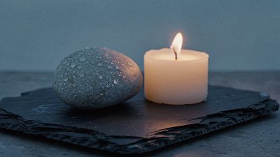 Зажжённая свеча рядом с обмытым водой речным камнем на тёмном сланце в сумеречном свете