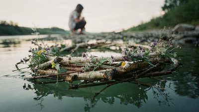 Девушка плетёт венок из берёзовых веток и полевых цветов у реки в солнечный день поздней весны